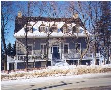 Front elevation of the Louis Bertrand House, showing the large, picturesque verandah with decorative balustrade encircling the house, and, underneath, the ground level gallery also enclosed by balustrade and gates, 1999.; Parks Canada Agency / Agence Parcs Canada, 1999