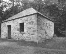 Corner view of the Blacksmith's Shop, showing the exterior walls, constructed of rough-faced masonry blocks, the limited number of windows and the door, 1989.; Public Works and Government Services Canada / Travaux publics et Services gouvernementaux Canada, 1989.