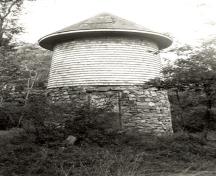 General view of the Water Tower, showing its tapered cylindrical shape, low-pitched conical roof, cedar-shingle cladding and rubble-stone base, 1992.; Parks Canada Agency / Agence Parcs Canada / Historica Resources Ltd., 1992.