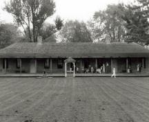 Front view of the Brick Barrack, showing its long rectangular bungalow structure with a prominent low hip roof which sweeps out to cover the long low verandah with its simple support posts, 1990.; Agence Parcs Canada, Bureau Régional de l'Ontario / Parks Canada Agency, Ontario Regional Office, 1990.
