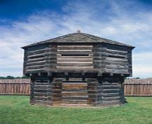 General view of the Octagonal Blockhouse showing the exterior walls constructed of squared logs with dovetailed corners, and the loopholes, 1978.; Agence Parcs Canada / Parks Canada Agency, T. Grant, 1978.