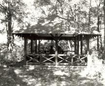 Side view of the Aubrey Island Picnic Shelter, showing the low surrounding railing of crossed timbers, 1992.; Archaeological Services and Historica Services, Ltd., 1992.