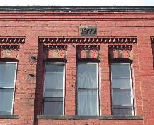This photograph shows one of the six triple set windows on the Germain Street façade, the modest cornice, decorative brickwork, and the 1877 datestone, 2004; City of Saint John
