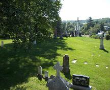 A photo of the tombstones of the Assomption Cemetery with the monument representing Jesus on the cross in the background; The Valley District Planning Commision