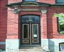 This photograph shows the entrance to the building, and illustrates the pedimented entablature, transom window, and detailed brick work, 2005; City of Saint John