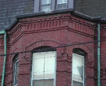 Cette photographie montre la corniche au-dessus de l'une des fenêtres en baie, les dentelets, les ouvertures à arche segmentée et le briquetage du fascia, 2005; City of Saint John