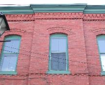 This photograph shows the central window above the entrance, the brick stringcourse under the sills and the brick corbel bands below the moulded wooden cornice, 2005; City of Saint John
