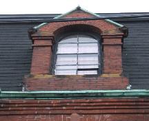 Cette image montre une lucarne à pignon en brique avec une fenêtre en bois à arc en plein-cintre. La corniche et les bandeaux d'encorbellement sont également évidents, 2005; City of Saint John