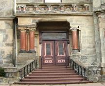 This image provides a view of the balustrade supported by Corinthian columns above the stone porch, the transom window and the paired wooden doors that are flanked by engaged Corinthian columns, 2005; City of Saint John